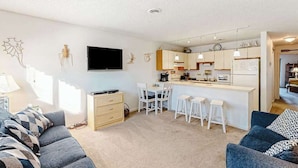 A modern living room with a sofa, a TV, a kitchen area with a bar, and a dining table with chairs. - Siesta Villa 304N (Ocean City)