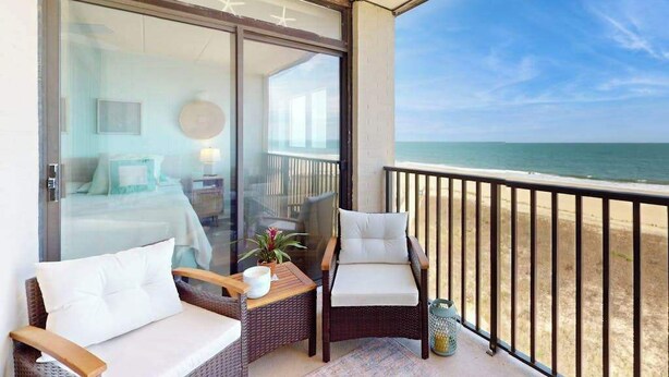 Un balcó amb moble de vimetes, vista a l'oceà i una porta de vidre que dona a una habitació amb un llit i un mirall rodó.
