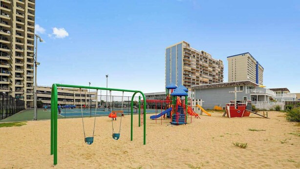 A playground with swings and slides, a small building, and high-rise buildings in the background. - Astounding beach condo with balcony, pools, gym, game room & tennis (Ocean City)
