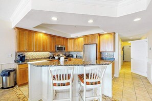 A modern kitchen with wooden cabinets, a stainless steel refrigerator, a microwave, and a granite countertop. - Belmont Towers 305 (Ocean City)