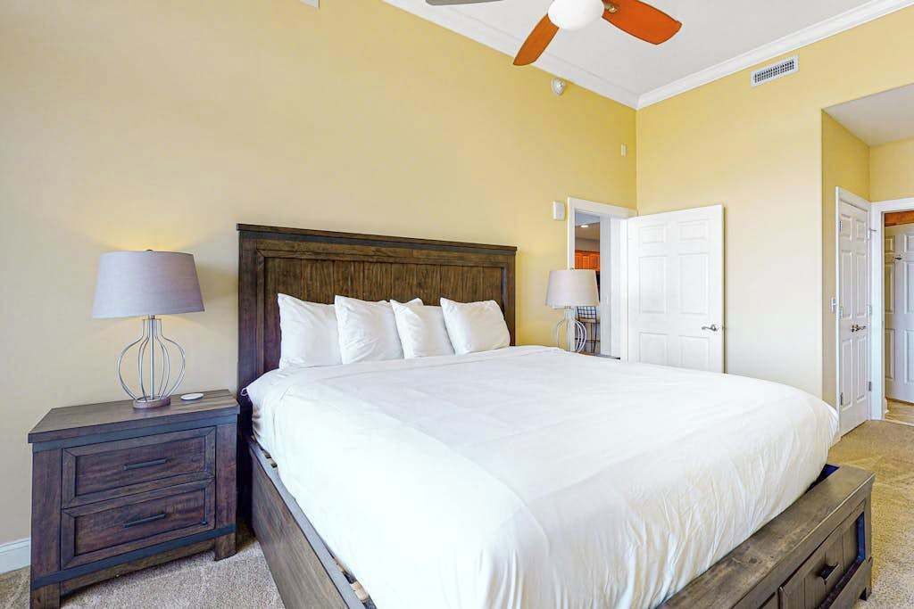 A bedroom with a large bed, a nightstand with a lamp, a ceiling fan, and a door leading to another room.