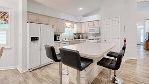 A modern kitchen with white cabinets, a central island with bar stools, a refrigerator, and a microwave. - Coral Shells (Ocean Pines)