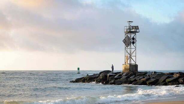 A lighthouse on a rocky outcrop by the sea, a sandy beach, and a person fishing. - Coral Shells (Ocean Pines)
