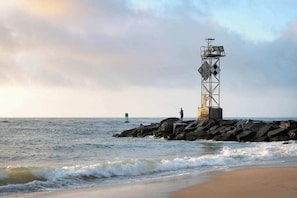 Ein Leuchtturm auf einem Felsen direkt an der Küste, ein Sandstrand und eine Person beim Angeln.