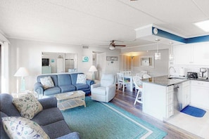 Ein modernes Wohnzimmer mit einer Couch, Sesseln und einem Couchtisch. Es gibt eine Küchenzeile mit weißen Schränken und einem Esstisch.