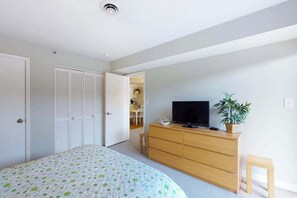 A bedroom with a bed, a TV on a wooden dresser, a potted plant, and a door leading to another room. - Well-Located Condo 2 Blocks from the Beach w/ AC & Private Laundry (Ocean City)