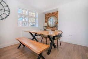 Een eethoek met een houten tafel, bankjes en stoelen. Er is een raam met meerdere ruitjes, een bakstenen muur en een ronde spiegel.