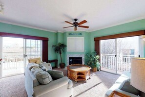 A living room with a sofa, a coffee table, a ceiling fan, a fireplace, and a balcony with a railing. - Bayside condo in Midtown with balcony, pool, sauna, gym & pier (Ocean City)