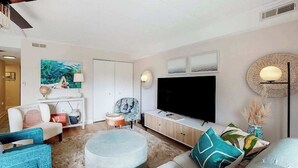 A modern living room with a flat-screen TV, a sofa, a round ottoman, and a coffee table. - Freshly Renovated Ocean Block Condo w/ Balcony, Partial Ocean View, AC & WiFi (Ocean City)