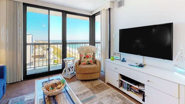 A modern living room with a large TV, a comfortable chair, a small table, and a view of the ocean through large windows. - Private beach condo with balcony, washer/dryer, pools, gym & tennis courts (Ocean City)
