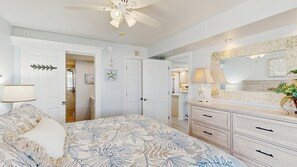 Una camera da letto con un letto, una cassettiera, uno specchio, una ventola a soffitto e una porta che dà su un'altra stanza.
