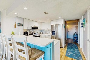 A modern kitchen with white cabinets, a central island, stainless steel appliances, and a blue patterned rug. - Unique beach condo near the water with balcony, pools, game room & AC (Ocean City)