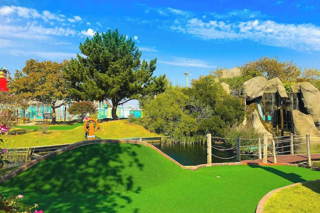 A miniature golf course with a green artificial turf, a pond, and a large tree.