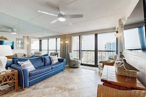 A spacious living area with a blue sofa, a dining table, and a balcony with a view. - Convenient & budget-friendly beach condo with balcony, pools, sauna & gym (Ocean City)