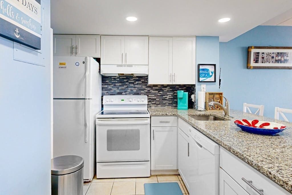 A kitchen with white cabinets, a stainless steel refrigerator, a built-in oven, a tiled backsplash, a granite countertop, a sink, and a framed picture on the wall.