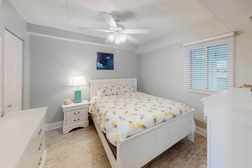A bedroom with a bed, nightstand, lamp, ceiling fan, window with blinds, and a wall-mounted picture.