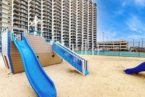 Un parque infantil con un tobogán azul y una estructura de madera, con un edificio residencial alto de fondo.