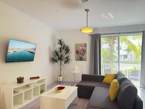 Ein modernes Wohnzimmer mit einem Flachbildfernseher, einer Couch, einem Couchtisch, einer Pflanze und einem großen Fenster mit Blick auf Palmen.