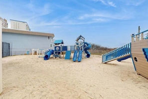 Ein Spielbereich im Freien mit Sand, einer Rutsche, Klettergerüsten und einer hausähnlichen Konstruktion.