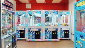 Eine Reihe von Prize Cube-Greifautomaten in einer hell erleuchteten Spielhalle mit rot-weiß karierter Decke.