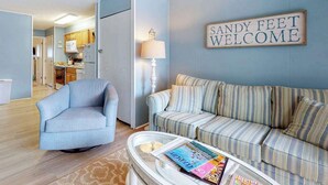 A living room with a sofa, a chair, a coffee table with magazines, a lamp, and a sign that reads 'SANDY FEET WELCOME'. - Sea Place 108 (Ocean City)