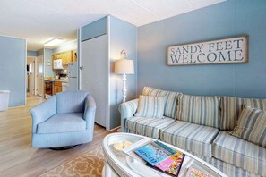 Ein Wohnzimmer mit einer Couch, einem Sessel, einem Couchtisch mit Zeitschriften, einer Lampe und einem Schild mit der Aufschrift 'SANDY FEET WELCOME'.