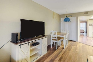 A living room with a flat-screen TV on a wooden stand, white chairs, a dining table, and a sign on the door. - Sea Mark 302 (Ocean City)
