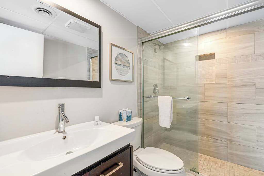 Une salle de bain avec un lavabo blanc, un miroir, un tableau encadré, des toilettes et une douche fermée par une paroi de verre.