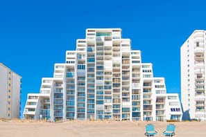 Un hôtel à plusieurs étages doté de balcons, avec un rez-de-chaussée recouvert de sable et un ciel d’un bleu limpide.