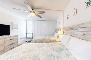 Une chambre à coucher avec un ventilateur de plafond, un lit doté d’une tête de lit en bois, un téléviseur et une fenêtre avec des stores.