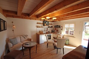 Ein gemütliches Wohnzimmer mit Sofa, Couchtisch und Sesseln, eine Küche mit weißen Geräten, Holzbalken und einem Fenster mit Blick.