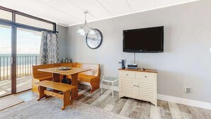 A room with a wooden dining table, a television mounted on the wall, and a view of the outdoors. - Oceanfront Condo w/ Great Beach View, Balcony, AC, W/D & Shared Pool/Tennis (Ocean City)