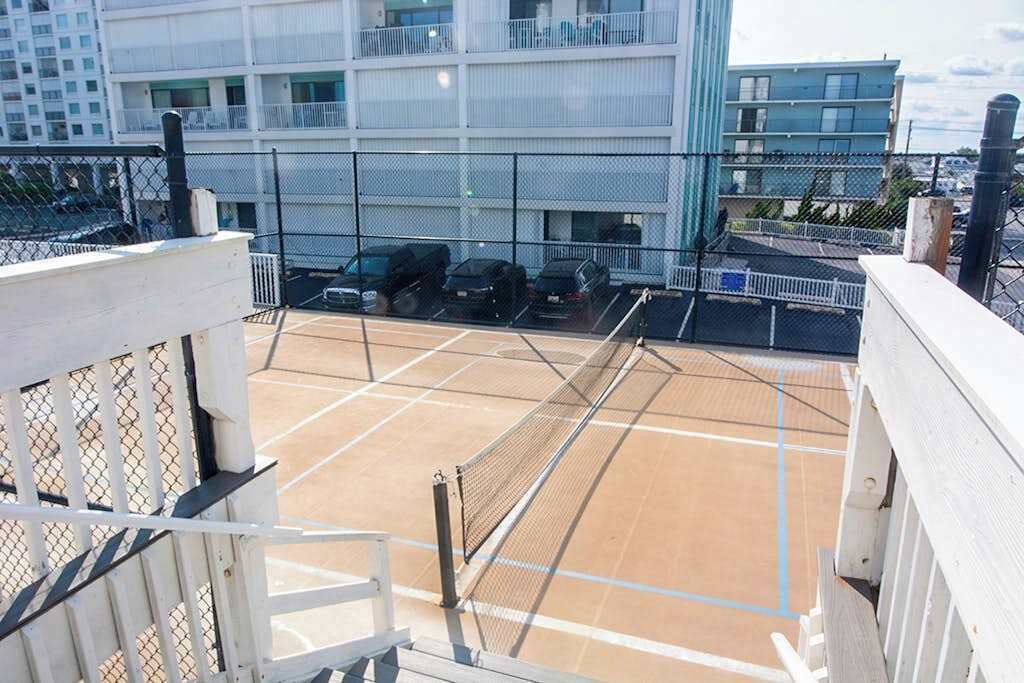 Una cancha de tenis con red, rodeada por una valla, y contigua a un edificio con balcones.