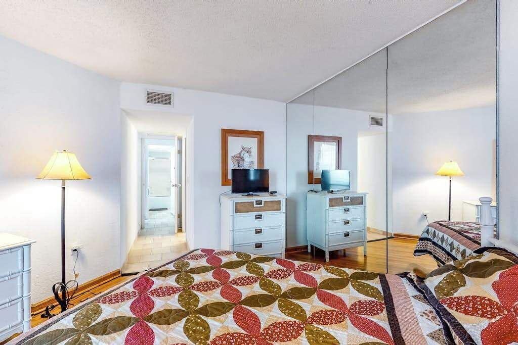 A bedroom with a bed, two bedside tables, a television, a mirror, and a door leading to another room.