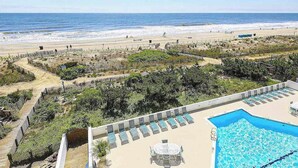 Zona de piscina con sillones y una mesa, con vista a una playa con cercado y vegetación.