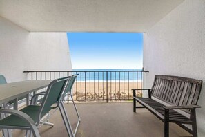 A balcony with a view of the beach, a table with chairs, and a wooden bench. - Oceanfront Condo with a Balcony, AC, WiFi, Ocean View & Shared Outdoor Pool (Ocean City)