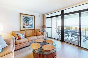 A living room with a glass coffee table, a sofa, and a large window with a view of outdoor furniture. - Oceanfront Condo with Covered Balcony, WiFi, AC, Laundry & Shared Pool (Ocean City)