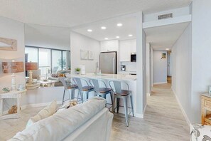A modern kitchen with white cabinets, a stainless steel refrigerator, and a bar area with bar stools. - Breezy, Oceanfront Condo with Balcony, AC, WiFi, Laundry & Shared Outdoor Pool (Ocean City)