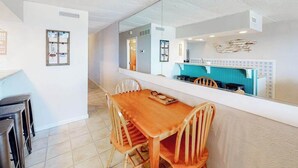 Een eetruimte met een houten tafel en stoelen, een grote spiegel die de keuken weerspiegelt, en een prikbord aan de muur.