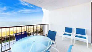 A balcony with a glass table and chairs, a view of the beach, and a white wall. - Newly Renovated, Oceanfront Condo with Balcony, Great View, AC & Shared Pool (Ocean City)