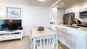 A modern kitchen with white cabinetry, a dining table with chairs, a flat-screen TV, and a ceiling light fixture. - Newly Renovated, Oceanfront Condo with Balcony, Great View, AC & Shared Pool (Ocean City)