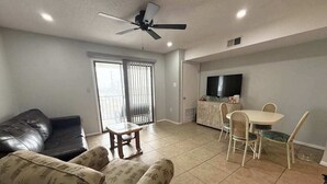 Ein Wohnzimmer mit einer Couch, einem Esstisch mit Stühlen, einem Deckenventilator und einem Fernseher an der Wand.