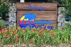 A wooden sign with the name 'Outrigger' and a colorful logo, surrounded by a garden with red flowers. - Outrigger 205 (Ocean City)