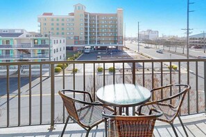 A balcony with wicker chairs and a glass table, overlooking a parking lot and a hotel building. - Outrigger 205 (Ocean City)