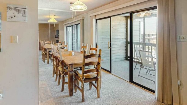 A dining area with a long wooden table and chairs, a sliding glass door leading to an outdoor balcony, and a ceiling light fixture. - Our Place At The Beach 306F (Ocean City)