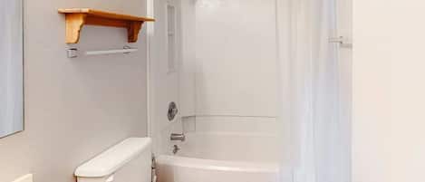 Et badeværelse med toilet, vask, badekar, bruseforhæng og en træhylde.