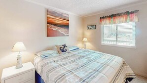 A bedroom with a bed, a nightstand, a lamp, a window with blinds, and a wall-mounted artwork. - Our Place At The Beach 306F (Ocean City)