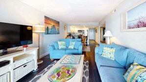 A living room with a TV, two blue sofas, a coffee table, and a kitchen in the background. - Renovated, Oceanfront Condo w/ Gorgeous Ocean Views, Balcony, AC & WiFi (Ocean City)