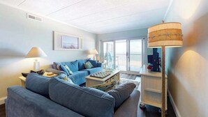 A living room with a sofa, a coffee table, a lamp, and a view of the outside. - Renovated, Oceanfront Condo w/ Gorgeous Ocean Views, Balcony, AC & WiFi (Ocean City)