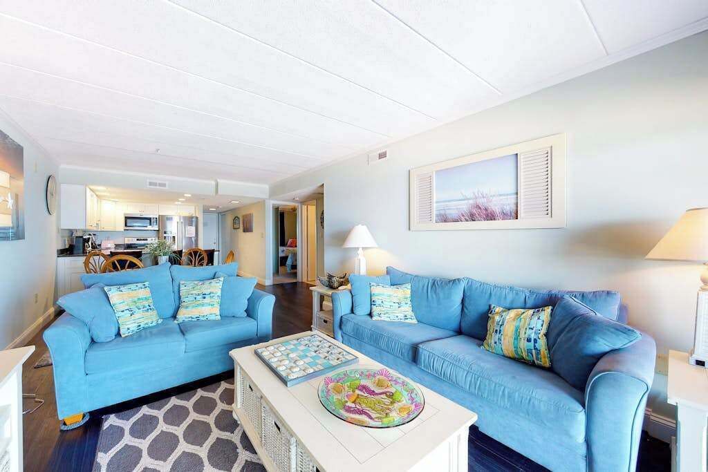 Ein Wohnzimmer mit zwei blauen Sofas, einem Couchtisch und einer Küche im Hintergrund.